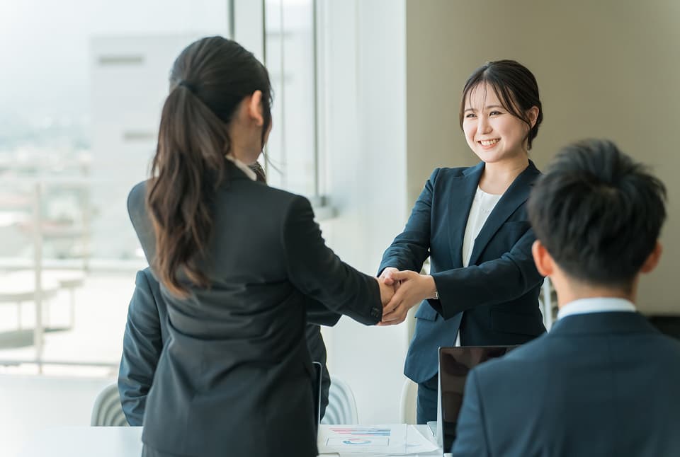 オフィスでスーツ姿の女性同士が笑顔で握手を交わし、商談の成功を感じさせる場面