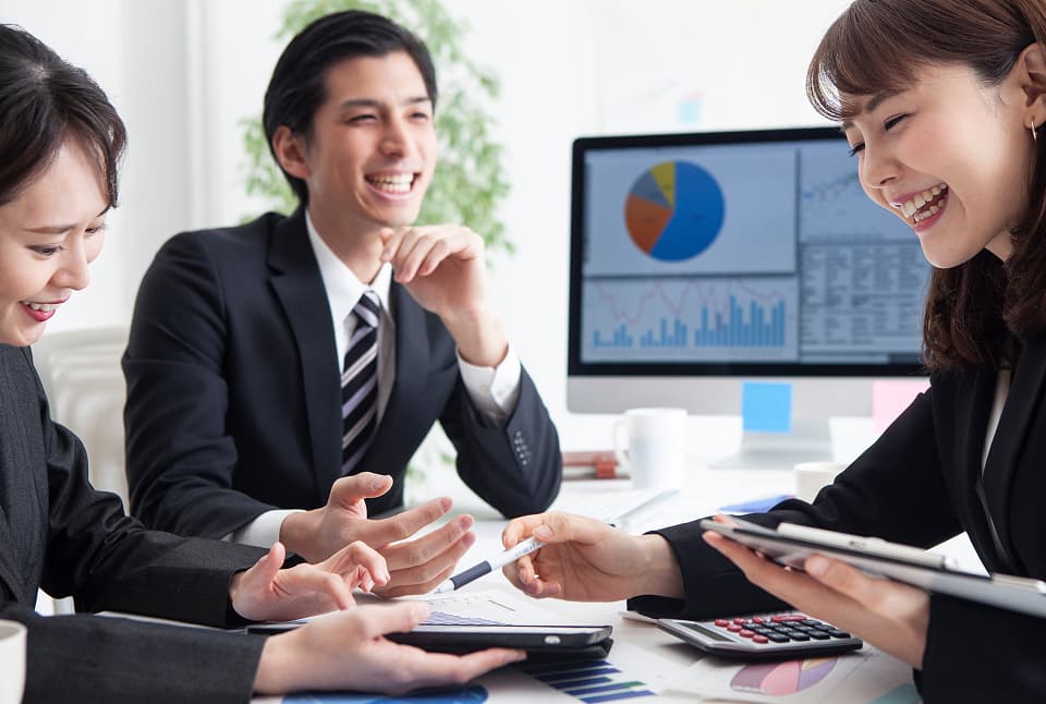 オフィスでスーツ姿の男女が笑顔で打ち合わせし、資料や電卓を使いながら話し合うビジネスシーン